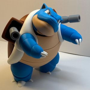 May include: A blue and tan creature figurine with a brown shell on its back, featuring two gray cannons extending from the shell. The figure has white claws, a white shell border, and a smooth, matte finish, standing in a bipedal, forward-leaning pose.