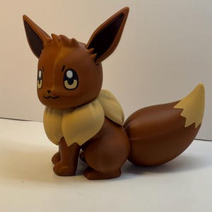 May include: A brown Eevee figurine with large pointed ears, dark brown inner ear details, and a cream-colored ruff around its neck. The character has large brown eyes, a small black nose, and a bushy brown tail with a cream-colored tip.