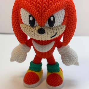 May include: A knitted figurine of Knuckles the Echidna, a character from the Sonic the Hedgehog franchise. The figure is primarily red with white, black, green, and yellow accents. The figure has a textured appearance, with visible knit stitches.