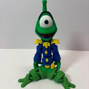 Puede incluir: Una figura articulada verde, azul y amarilla de un extraterrestre de dibujos animados con un ojo grande. La figura tiene una cabeza verde con una antena negra, un ojo blanco con una pupila negra y una boca roja. Viste una chaqueta azul con charreteras y botones amarillos.