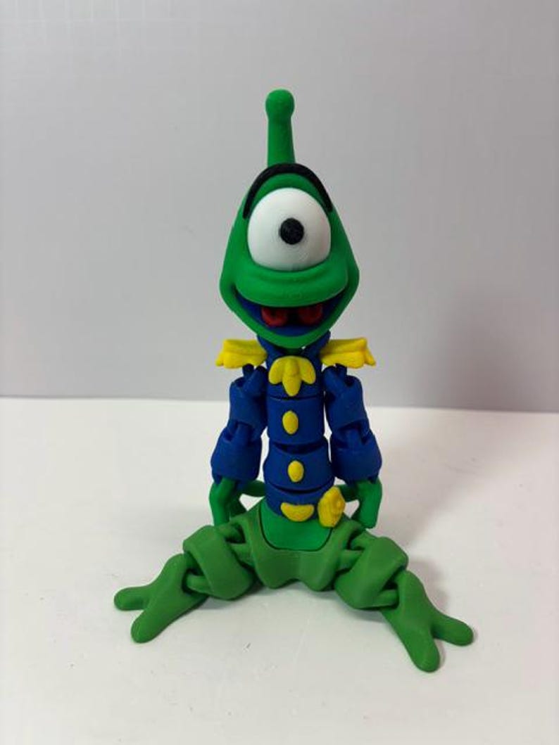 Puede incluir: Una figura articulada verde, azul y amarilla de un extraterrestre de dibujos animados con un ojo grande. La figura tiene una cabeza verde con una antena negra, un ojo blanco con una pupila negra y una boca roja. Viste una chaqueta azul con charreteras y botones amarillos.