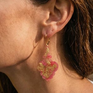May include: A pink and gold earring is shown close up. The earring has a gold-coloured hook and a pink, heart-shaped pendant with gold filigree. The earring is hanging from an ear.