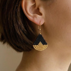 May include: Gold-coloured earrings with a triangular design. The top section is black, and the bottom is gold, made of small beads. The earrings are suspended from a gold hook.