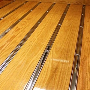 May include: Close-up of a wooden truck bed floor with parallel, polished chrome strips. The wood has a warm, golden-brown hue, and the metal strips are secured with small, silver rivets. The design is a classic, vintage style.