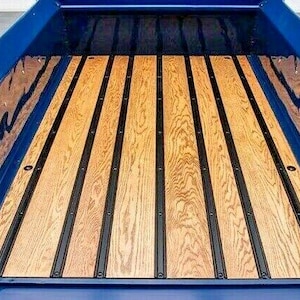 May include: A close-up view of a truck bed with a wooden floor. The floor is made of light brown wooden planks with dark brown metal strips running between them. The truck bed is painted a glossy blue.
