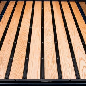 May include: Close-up of a truck bed with light brown wooden planks and black metal strips. The planks run lengthwise, creating a textured surface. The black strips are evenly spaced, adding a contrasting element to the design.