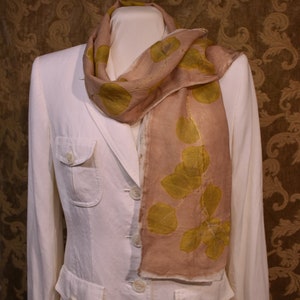 Peut inclure: Écharpe beige avec un motif de feuilles dorées et vertes, drapée sur un blazer blanc. L'écharpe est faite d'un tissu léger et présente un motif de feuilles répétitif. Le blazer a un design classique avec des boutons et des poches.