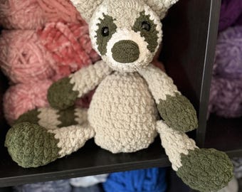 May include: A handmade crochet raccoon plush toy with a gray and white body, green accents, and black eyes. The plush is sitting on a black shelf with various colors of yarn in the background.