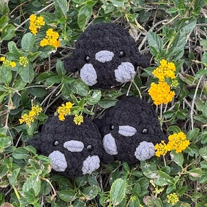 May include: Three handmade black crochet orca plushies with gray accents, nestled among green foliage and bright yellow flowers. The plushies have small black eyes and gray patches.