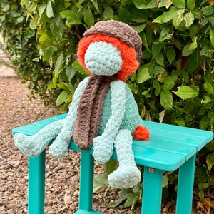 May include: A handmade crochet doll with a teal body, brown hat, and scarf, and orange accents. The doll is sitting on a teal bench. The doll's features include a fluffy orange trim around the face and a small orange pom-pom.