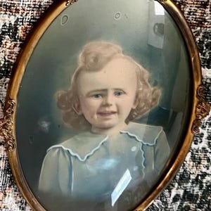 May include: An antique oval portrait of a child in a gold ornate frame. The portrait features a young child with curly blonde hair and a light blue dress. The frame has decorative detailing.