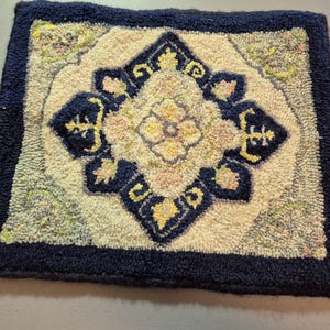 May include: A square, hand-hooked rug with a navy blue border. The rug features a central diamond design in cream, navy, yellow, and pink. The design has a floral motif. The rug is likely made of wool.
