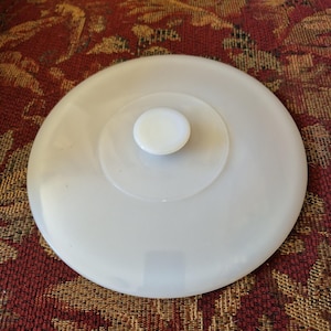 May include: A white, round lid with a central knob. The lid has a layered design with concentric circles. The knob is a small, rounded shape. The lid is made of a smooth, opaque material.