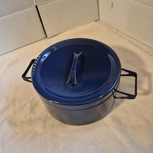 May include: A navy blue enameled cast iron Dutch oven with a matching lid and black metal handles. The pot is round and has a handle on the lid. The Dutch oven is sitting on a beige surface.