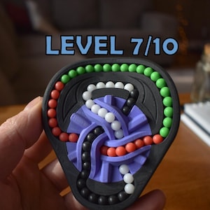 Könnte beinhalten: Ein schwarzes Puzzlespielzeug mit bunten Perlen und dem Text "LEVEL 7/10". Das Puzzle hat eine lila Mitte und Bahnen, durch die sich die Perlen bewegen können. Die Perlen sind rot, grün, weiß und schwarz.
