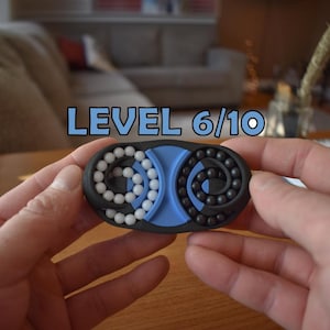 May include: A black, blue, and white puzzle toy with the text "LEVEL 6/10". The toy has two sections, one with white beads and the other with black beads. The puzzle is held in two hands.