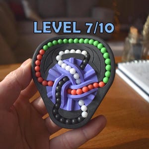 Peut inclure: Un casse-tête triangulaire noir portant l'inscription LEVEL 7/10. Le puzzle comprend une structure centrale violette avec des pistes entrelacées contenant de petites perles sphériques blanches, noires, rouges et vertes disposées en chemins.