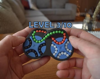 Pode incluir: Um quebra-cabeça de plástico preto em forma de oito com o texto LEVEL 3/10. O jogo contém pequenas esferas nas cores verde, laranja, preta e branca dispostas em trilhas circulares azuis inseridas na estrutura preta do objeto.