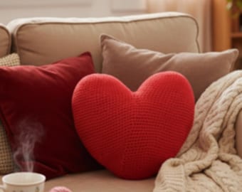 Puede incluir: Una acogedora sala de estar con un sofá beige adornado con cojines rojos y beiges. Una gran almohada roja en forma de corazón, tejida a ganchillo, es el punto focal. Una taza de té humeante sobre una mesa auxiliar con lana y una manta de punto.