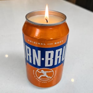 May include: A lit candle in an orange and blue IRN-BRU can. The can features the words "ORIGINAL and BEST" and "IRN-BRU". The bottom of the can says "MADE IN SCOTLAND FROM GINGER".