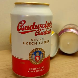 May include: A white Budweiser beer can with red and gold detailing. The can displays the Budweiser logo in red, the words "ORIGINAL CZECH LAGER", and a crest. The base of the can is red, with the text "OWNED BY THE CZECH REPUBLIC".