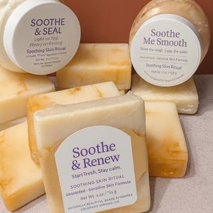 May include: An assortment of skincare products, including jars and bars of soap. The jars have white lids with text that reads "Soothe & Seal" and "Soothe Me Smooth." The soap bars are rectangular and wrapped in tan paper with the text "Soothe & Renew."
