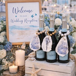 May include: A wedding reception display with three Prosecco bottles, each with seashell tags and wooden bead necklaces. A sign reads "Welcome to our Wedding Reception". The scene includes candles, flowers, and a starfish, all on a sandy surface.