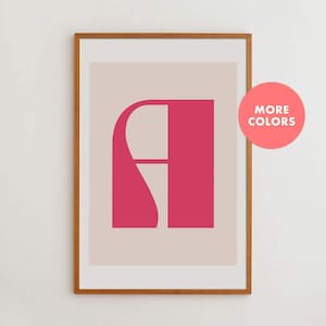 May include: A framed wall art print featuring a stylized, bold magenta letter A centered on a light beige background. The wooden frame is a medium brown tone. A circular pink badge in the upper right corner contains the white text MORE COLORS.