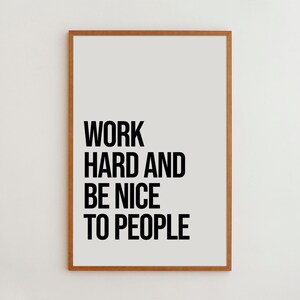 May include: A rectangular wall art print in a light brown wooden frame featuring the black text WORK HARD AND BE NICE TO PEOPLE centered on a plain off-white background. The typography is bold, sans-serif, and arranged in four distinct lines.