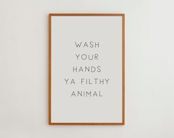 May include: A rectangular wall art print in a medium-toned wooden frame. The print features a light gray background with black sans-serif text that reads: WASH YOUR HANDS YA FILTHY ANIMAL. The design is centered and occupies the majority of the frame.