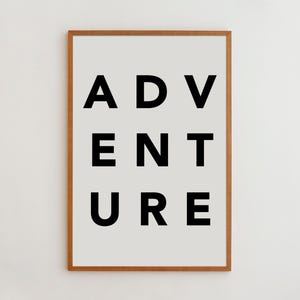 May include: A rectangular wall art print in a light brown wooden frame. The print features a light gray background with the word ADVENTURE printed in bold, black, sans-serif capital letters, arranged in three rows of three letters each.