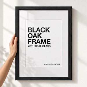 May include: A black oak picture frame with a white mat and clear glass. The white insert features black text that reads: BLACK OAK FRAME WITH REAL GLASS, and in the bottom right corner, the text: Crafted in the USA. The frame has a wood grain.