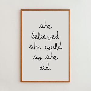 May include: A rectangular wall art print in a light brown wooden frame. The print features a light gray background with the black handwritten-style text: she believed she could so she did. The design is centered within the frame.