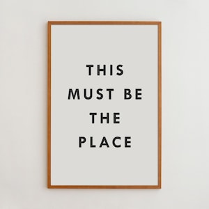 May include: A rectangular wall art print featuring a light gray background and a thin wooden frame. The text THIS MUST BE THE PLACE is printed in bold, black, sans-serif capital letters centered in the middle of the print, creating a clear visual.