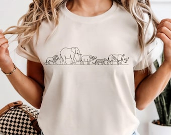 Puede incluir: Una camiseta de manga corta color crema con un gráfico de líneas negras en el pecho. El diseño muestra una fila horizontal de cinco animales de safari africanos, incluyendo un león, un elefante, un búfalo, un leopardo y un rinoceronte.