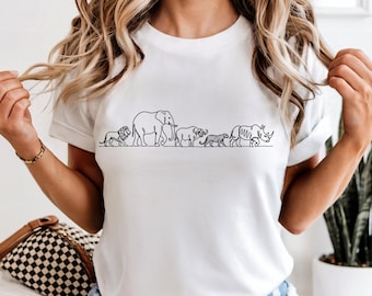 May include: A white short-sleeved t-shirt featuring a black line art graphic of five African safari animals walking in a single file line. The design depicts a lion, an elephant, a buffalo, a leopard, and a rhinoceros on a plain white background.