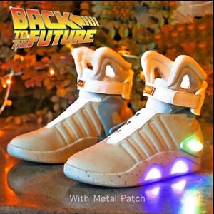 May include: Pair of futuristic-style sneakers with the text "BACK TO THE FUTURE". The shoes are beige and white with a light-up sole and accents. The shoes have a strap and a metal patch.