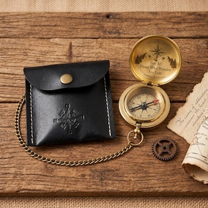 May include: A brass pocket compass with a hinged lid featuring the text "Not All Those Who Wander Are Lost" and a mountain graphic, attached to a gold-tone chain and a black leather pouch with a snap button and embossed compass rose design.