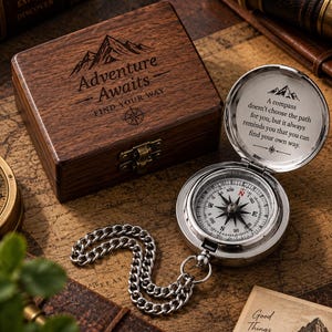 Può includere: Una bussola da tasca argentata con catena e una scatola in legno scuro. Il coperchio presenta un'incisione di una montagna e il testo Adventure Awaits FIND YOUR WAY. L'interno del coperchio riporta il testo A compass doesn't choose the path for you, but it always reminds you that you can find your own way.