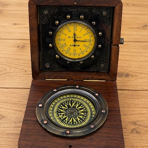 May include: A dark brown wooden box containing a clock and a compass. The clock has a yellow face with Roman numerals and the text 49. Bond Street LONDON. The compass features a yellow dial with black markings and a brass-toned metal rim.