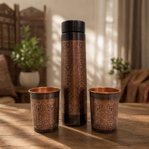 Peut inclure: Une grande bouteille d'eau cylindrique en cuivre avec un bouchon noir et deux gobelets en cuivre assortis. Chaque article présente une finition brun foncé avec un motif floral damassé répétitif et complexe en cuivre métallique.