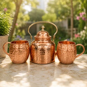 May include: A polished copper tea set consisting of a central kettle with a rounded lid and a curved handle, flanked by two matching mugs. Each piece features a distinct hammered texture across the exterior surface, reflecting light with a warm glow.