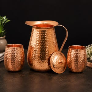 May include: A copper pitcher with a hammered texture, a curved handle, and a matching lid placed beside it, accompanied by two matching hammered copper tumblers. The set features a polished metallic finish with a distinct dimpled surface pattern.
