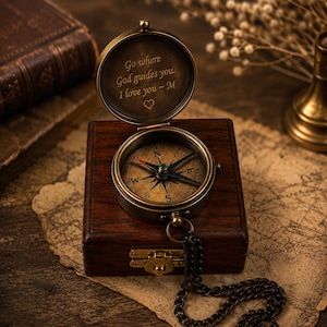 Può includere: Una bussola da tasca in ottone con coperchio incernierato recante il testo inciso 'Go where God guides you. I love you ~ M' e un simbolo a forma di cuore. La bussola poggia su una scatola di legno scuro con chiusura in metallo e catena.