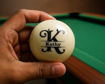 May include: A cream-colored billiard ball with the name "Kathy" and a stylized "K" in black. The ball is held in a hand, with a green pool table in the background.