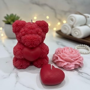 May include: A selection of decorative candles: a red teddy bear, a pink rose, and a red heart. The teddy bear candle is covered in rose-shaped details and has a heart on its chest. The rose candle is pink, and the heart candle is red.