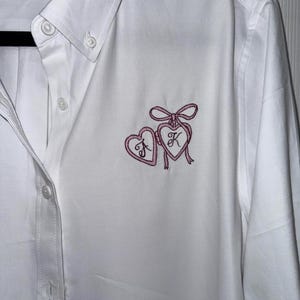 May include: White button-down shirt with a pink embroidered design of two hearts and a bow. The hearts contain the letters "J" and "K". The shirt has a classic collar and buttoned cuffs.