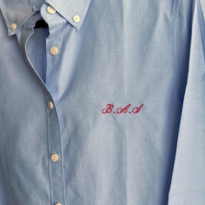 May include: Light blue button-down shirt with a classic collar and long sleeves. The shirt features white buttons and red embroidered initials "B.A.S" on the chest. The fabric has a subtle texture, and the shirt is neatly pressed.