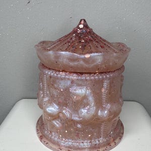 Op de afbeelding: Een roze pot met een carrouselthema en een bijpassend deksel. De pot is cilindrisch met reliëf carrouselpaarden en bedekt met glitter. Het deksel is conisch en ook bedekt met glitter. De pot staat op een wit oppervlak.