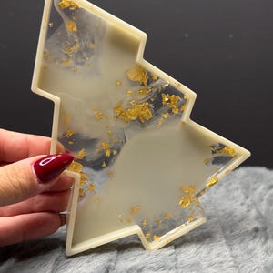 Könnte beinhalten: Ein weihnachtsbaumförmiges Tablett aus cremeweißem Harz mit eingebetteten Blattgoldflocken. Das Tablett hat eine glatte, glänzende Oberfläche mit wirbelnden Mustern aus undurchsichtigem und durchscheinendem Harz.